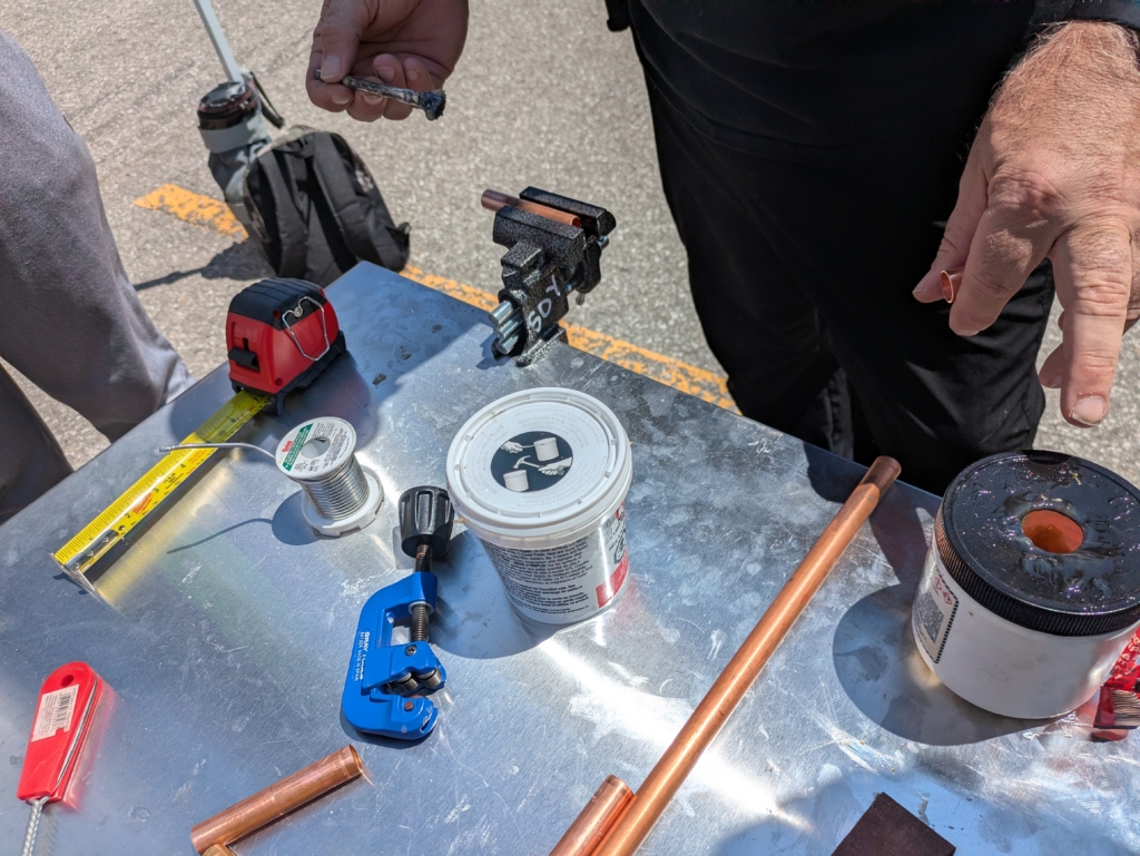 Tools for working with copper pipes including clamps, measuring tape, soldering wire (the spool of wire), pipe cutter (the small blue thing in the middle), a plastic jar of adhesive (referred to as Flux, on the right with a hole drilled into the top of the lid), emery cloth and wire brush for removing oxidation.